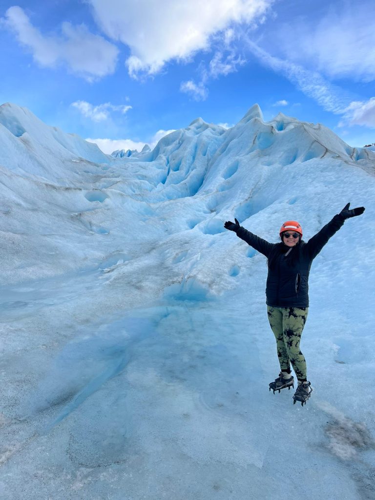 A woman wearing a helmet and crampons with her arms in the air, stood on the Perito Moreno glacier in Argentina